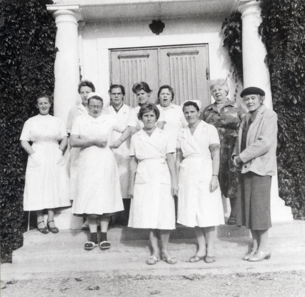 Elsa Tiberg, kallad "Fröken",  klädd i mörk hatt med del av sin personal på trappan vid Bällsta gård - Vallbostrands vårdhem. Elsa Tiberg hade först pensionat på gården, 1928-1933 och sedan vårdhem för vuxna med utvecklingsstörning.  F v står Carla Lilja som var husmor de sista 10 åren, Fru Johansson, okänd, Svea Gullmes,    Med på bilden är också Helga som jobbade i köket och Ester Granberg (med vit mössa) som arbetade som tvätterska. Margareta Bertilsson står bredvid Elsa Tiberg  Vårdhemmet hade ca 13 anställda.  Jfr 90 04 00: 003