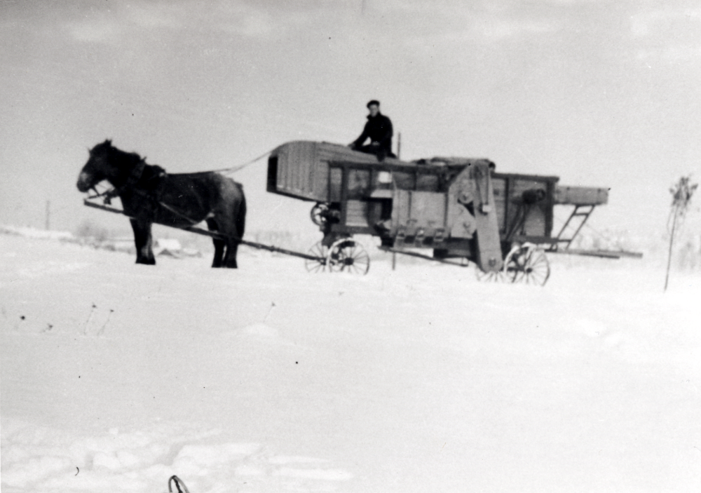 Hästdraget tröskverk på väg mot Prästgården.  På tröskan sitter Harald Ek.  Tröskan drevs av en speciell motordel, som kopplades med remdirft till tröskans drivhjul.  I bakgrunden skymtar skorstenen från Vallentuna tegelbruk.  Hästan och tröskan har just passerat järnvägen på gamla vägen mellan Vallentuna kyrka och Klockargården.  I förgrunden syns Josef Anderssons skidstav som han slängt i från sig före fotograferingen