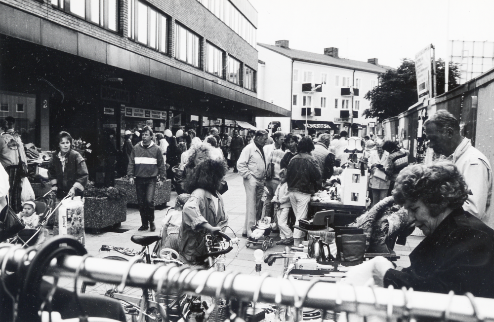 Vallentuna marknad på Tuna torg den andra lördagen i september, arrangerad av Vallentuna hembygdsförening. Här syns människor samlade runt de olika stånden. I förgrunden hänger en stång med kläder, endast galgkrokarna syns. T h ett plank uppsatt under byggnationen av det nya kommunalhuset