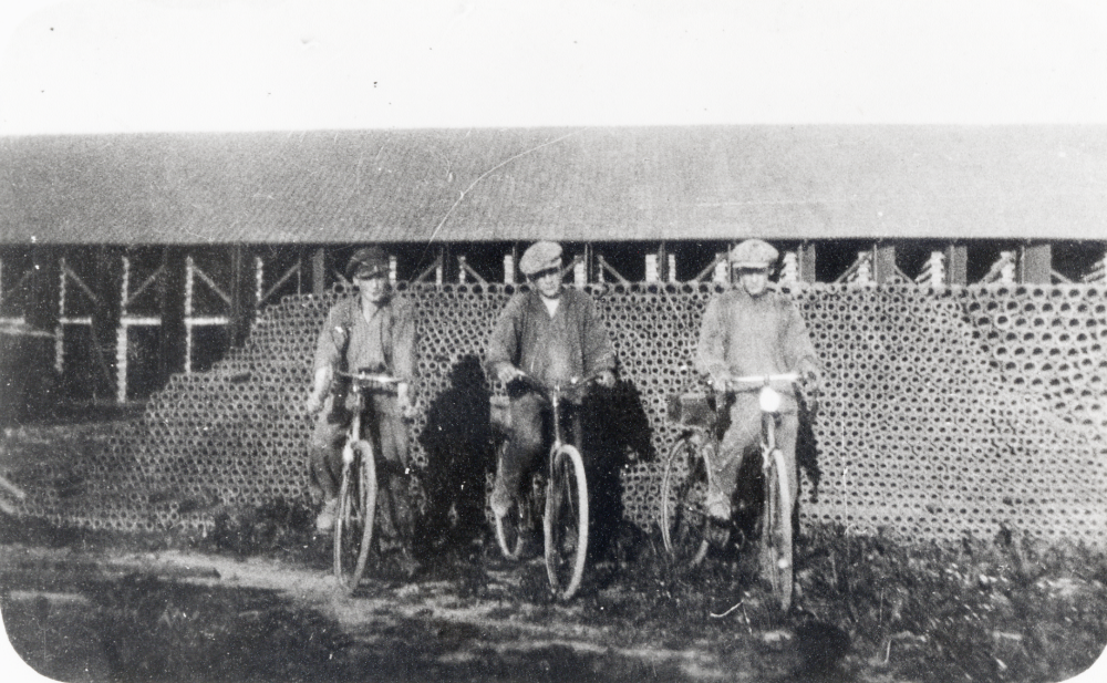 Åby tegelbruk.  Framför ett lager av tegelrör i två olika dimensioner  står fr v Max Karlsson, Sven Nordström och Harry Skogberg.  Alla tre sitter på cyklar.  På pakethållaren ser man unikaboxar - innehållet kunde vara termos med kaffe och smörgås för matrasten