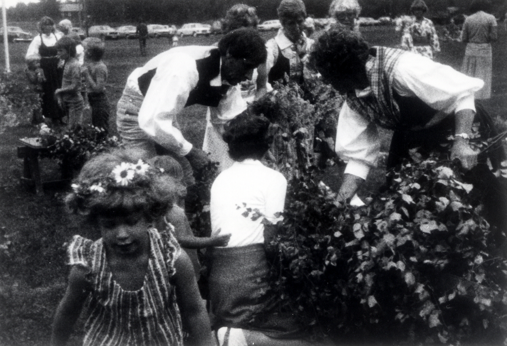 Midsommar firas på Björkvallen i Frösunda. Här lövas midsommarstången. I förgrunden syns Malin Hellsten med blomsterkrans i håret. Vid midsommarstången står fr v Inge Olsson, skymd okänd, Lillian Olsson, okänd dam i glasögon och Ulla Öhrlund, sittande på huk Ulla-Britt Helgöstam. Gruppen t v är fr v Andreas Jansson, Gunilla Jansson, okänd, Robert Roos och Joakim Jansson