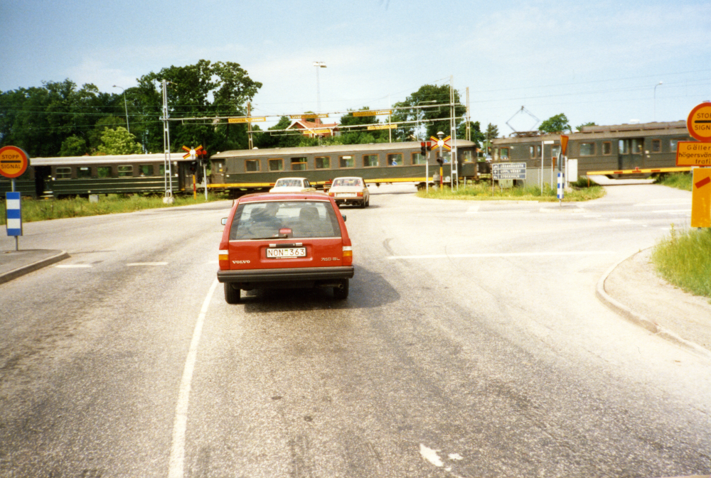Järnvägskorsningen söder om Vallentuna station kallades ofta ”Röda korset” för att det ofta blev stopp och långa köer där innan den planskilda korsningen byggdes. Här syns bilar som stannat till på Angarnsvägen framför nedfällda bommar och ett tåg som passerar på Roslagsbanan.
