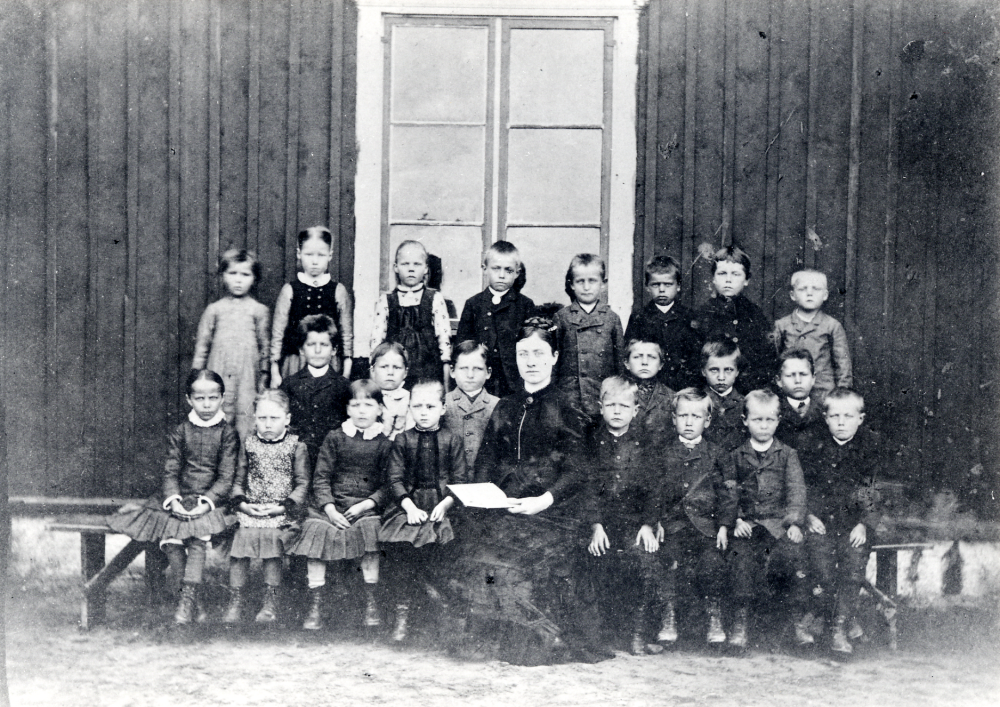 Lärarinna Ada Edlund tillsammans med barnen vid Björkholmens baptistkapell i Kårsta. Kårsta fria folkskola drevs i baptisternas regi 1861-1890, då skolan förbjöds. Ada Edlund tjänstgjorde i skolan 1870-1890. Undervisningen skedde först i en ambulerande skola i olika gårdar, ett skolhus och kapell byggdes klart 1863. 1864 byggdes ett internat för barn bosatta långt bort.