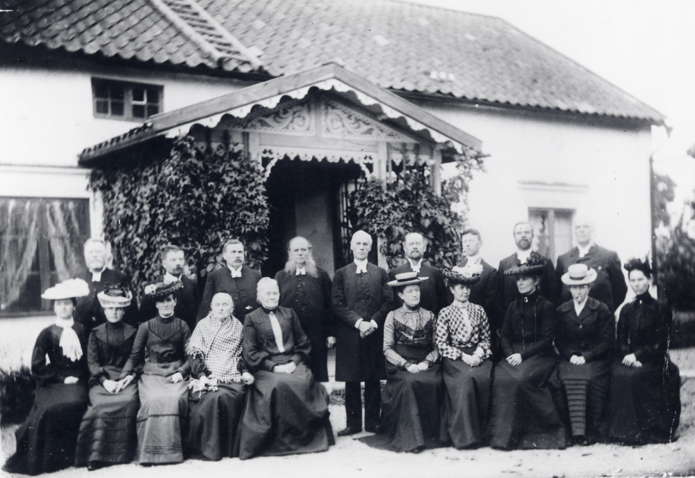 Kyrkoherde Carl Mauritz Anjou med vita polisonger i bildens mitt utanför Frösunda prästgård vid ett prästmöte omkring år 1900. Sittande nr 2 fr v är fotografens fru Gunhild Carlsson-Waldebäck