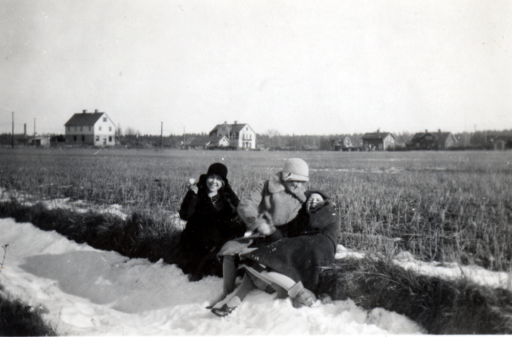 Tre flickor på vägen över Prästgårdsgärdet. Fr v en syster Svedberg, Rut Gustafsson, dotter till skomakare K P Gustafsson i Villa Edsberg, samt ytterligare en syster Svedberg.  I bakgrunden längs Allévägen syns fr v Ångbageriet, Valfrid Jonssons affär (senareTibells affär) och Vallentuna järnvägsstation