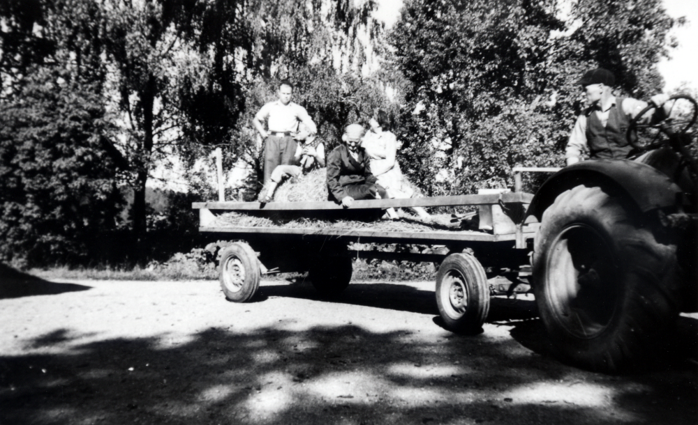 På gårdsplanen vid Ekskogens gård.  I brist på bil får man ta traktorn på utfärden. Traktorn drar en f d bryggerivagn med gummihjul.  Manfred Ekholm sitter vid ratten. På flaket sitter passagerna på höbalar fr v Bertil Sundmark, Timo Ekholm, Inez Ekholm och Vera Sundmark.  Inez och Manfred Ekholm ägde Ekskogen gård 1947-1965, Familjen Sundmark hyrde gårdens övervåning sommartid.

Timo och hans lillebror Sami kom till Sverige som finska krigsbarn 1944. Timo adopterades senare av ägarparet på Ekskogens gård i Kårsta, Inez och Manfred Ekholm. Läs mer i Timo Ekholms bok Krigsbarn 40.