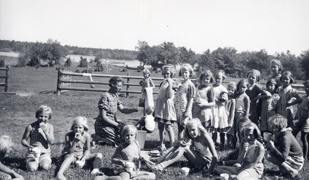 Margareta Andersson gift med Josef Andersson serverar flickorna mellanmål vid Barnens dags koloni i Tranvik på Singö i Stockholms norra skärgård. Margareta var tre somrar i rad biträde på kolonin. éven Josef och barnen Jerker och Svante vistades på kolonin. Vid måltiderna fick inte flickor och pojkar äta tillsammans