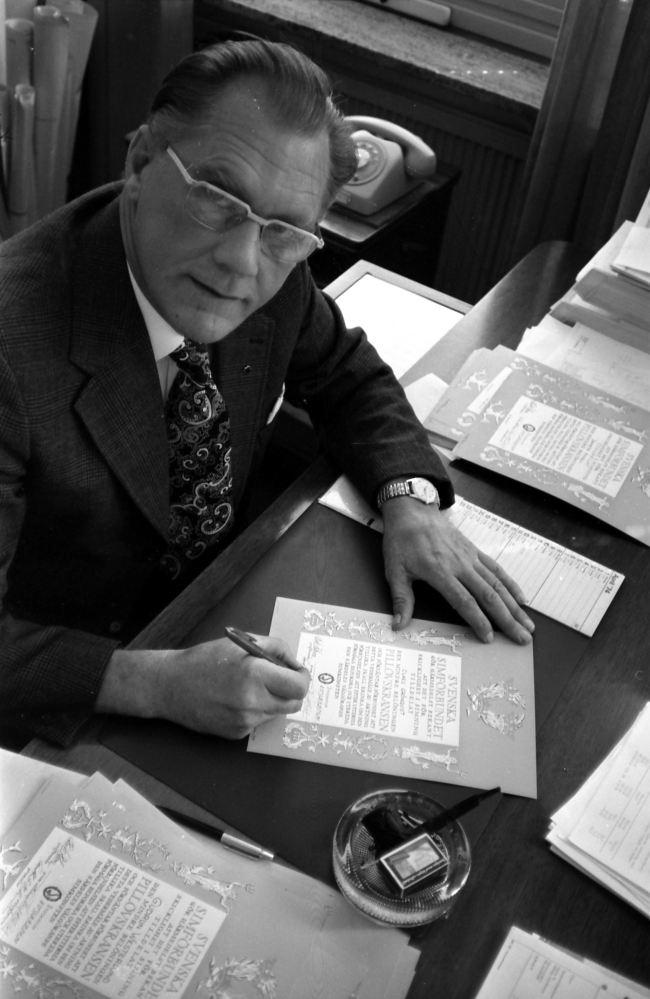 Kommunalrådet Karl-Erik Wikström (C) på sitt kontor signerar diplomen med Pillövskransen från Svenska Simförbundet som tilldelas deltagarna för "skicklighet i simning".
•Fotografiet ingår i bildserien ”Vardagsliv i Vallentuna kommun” av Rachael Gough-Azmier & Gunilla Blomé, Haga Studios HB.
