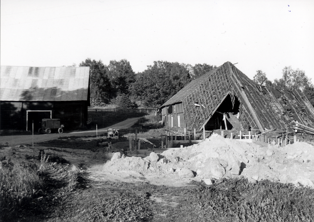 Ormstavägen från väster med Ormsta gårds lada t v och ladugård t h strax före rivningen.  Mellan husen skymtar Schramsta.  Ladugården är redan delvis riven. Bottenplattan har gjutits till ett av de första husen på Tegelvägen