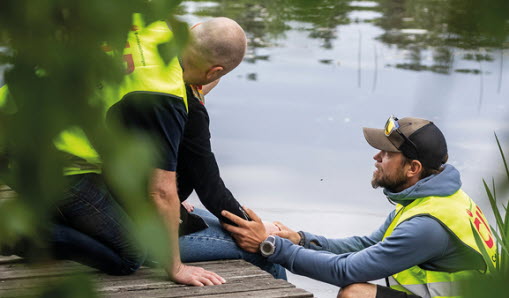 Två civila insatspersoner hjälper en person som sitter på en brygga vid vattnet.