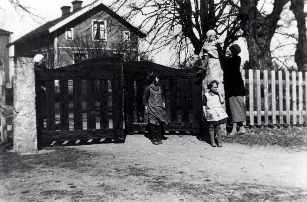 Syskonen Karlsson, Marianne, Ulla och lilla Sven-Erik uppe  vid grindarna till Väsby gård i Vallentuna.  Barnens föräldrar är Märta och Sven Karlsson, arrendator vid Väsby gård från 1930 till 1963.  Den goda vännen i familjen, fröken Lindblad håller i lille Sven-Erik, som sitter uppe på ena grindstolpen.  Bakom grinden står barnens mor Märta