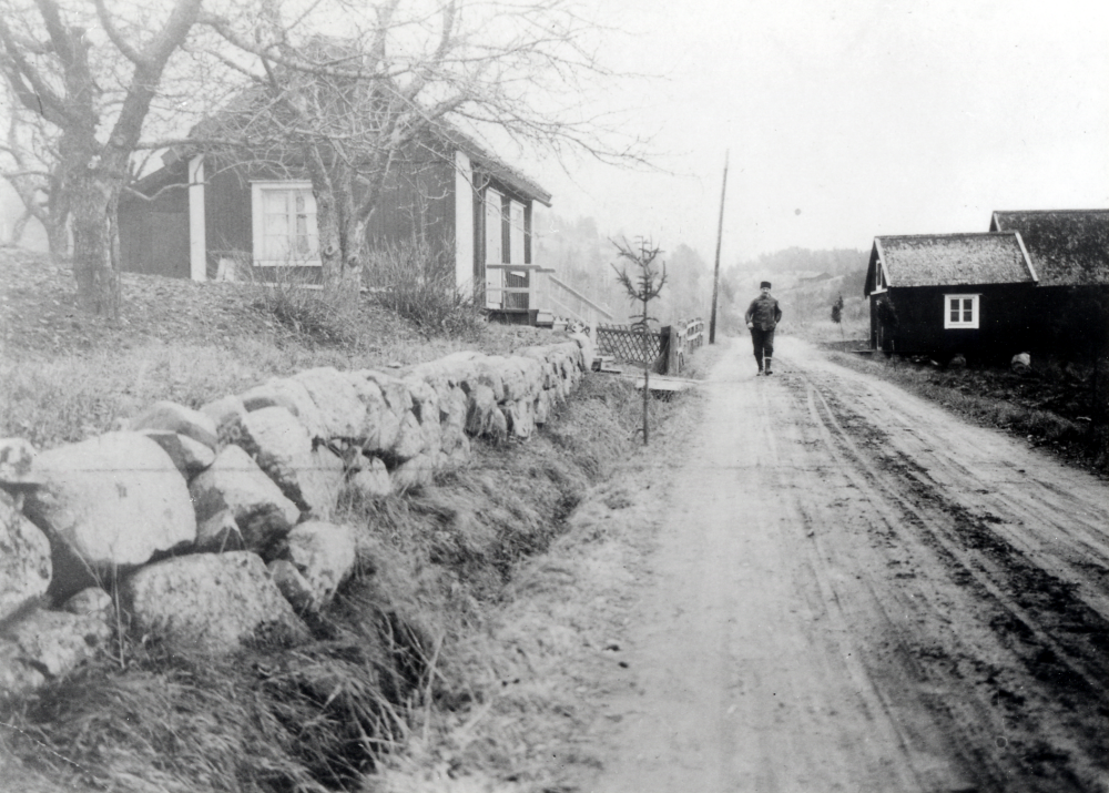 Klockarborg, "Söderbergarnas" i Lilla Garn. Namnet Klockarborg myntades av Harald "Halle" Söderberg i mitten av 1930-talet. Haralde bodde på Klockarborg tillsammans med sina syskon. På vägen, som kommer från Össeby-Garns kyrka och går mot Österåker, går Johan Jäger Lundberg boende på hemmanet Backebo-Lugnet. I bakgrunden skymtas Garnsberget och taket till torpet Frosenberg. Längs vägen är snöruskor uppsatta inför den kommande vintern. Till vänster i bilden syns Klockarborg med ett så kallat Jägarstaket mot vägen. Till höger syns Söderbergarnas fiskestugor.
Original och uppgifter John-Lars de Chasseur.