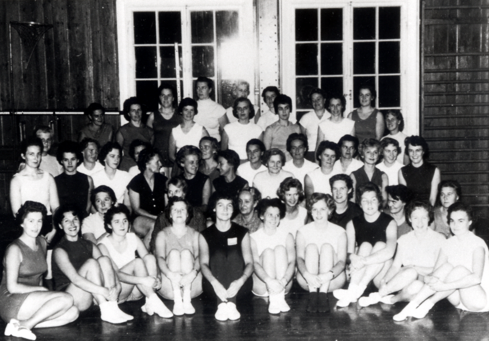 Husmodersgymnastiken i Vallentuna, grupperna I och II i Konsum Väsbys regi, förmodligen samlade i Röda skolans gymnastiksal i Hjälmsta.  Ledare för barn- och husmodersgymnastiken var Kerstin Andemark, som sitter i mitten klädd i mörk gymnastikdräkt.  Se Konsum i Norrort 1907-1997, s 114