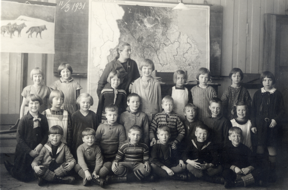 Lärarinnan Anna Gauffin tillsammans med skolbarnen inne i Hjälmsta skola Stående längst bak: Lille-Mor Karlsson, Elsa Öjdahl, Gunvor Pettersson, Margit Hedlund, Margit Grandin, Karin Wallberg, Eva Andersson, och Irma Jansson. Knästående fr v: Gunilla Härje, Anna-Lisa Johannesson, Ingegerd Gustavsson, Arne Andersson, Ragnar Pettersson, Birger Noaksson, Hjalmar Olsson, Gösta Ahlin, och Eva Åkerberg. Sittande längst fram fr v: Ove Karlsson, Olle Andersson, Erik Mörk, Folke Wallberg, Arne Jansson, och Rune Eriksson