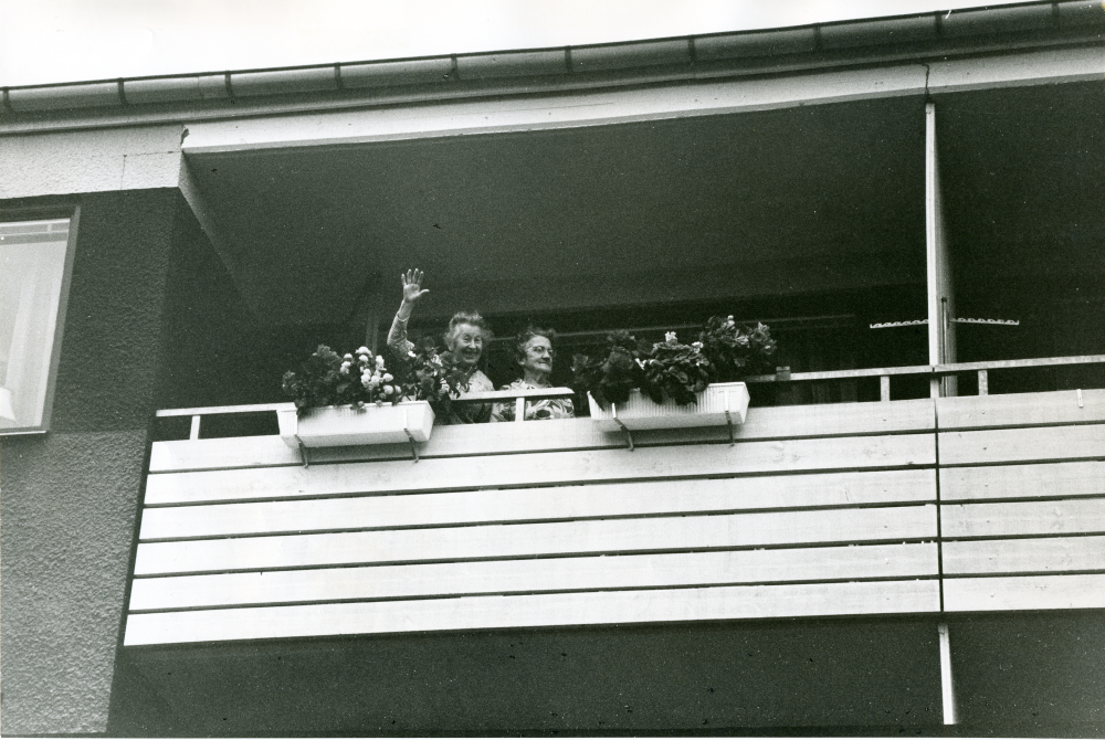 Gårdsfest i bostadsrättsföreningen Granen med bostäder på Fornminnesvägen och Teknikvägen. Två kvinnor deltar på avstånd och vinkar från en balkong.
•Fotografiet ingår i bildserien ”Vardagsliv i Vallentuna kommun” av Rachael Gough-Azmier & Gunilla Blomé, Haga Studios HB.
