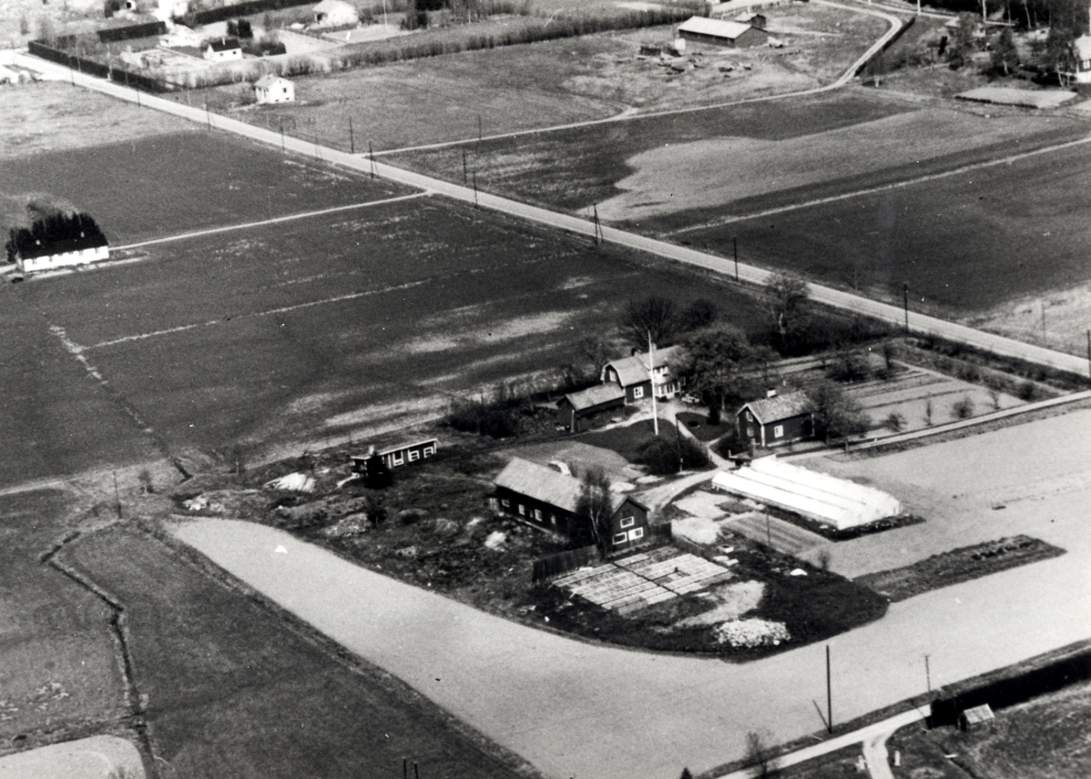 Flygfoto över Slumsta handelsträdgård i Lindholmen.  Agnes och Folke Hallgren köpte Slumsta gård 1945.  Trots  hård kyla och snö påbörjades det första växthuset 1946 - det hus som ännu står kvar 2000.  Agnes Hallgren körde bil tidigt på morgonen in till Kornhamnstorg i Stockholm med trädgårdens produkter. På kvällen skulle nästa dags transport förberedas.  T v syns "Gula Längan"  Stenby bostad för familjen Jansson