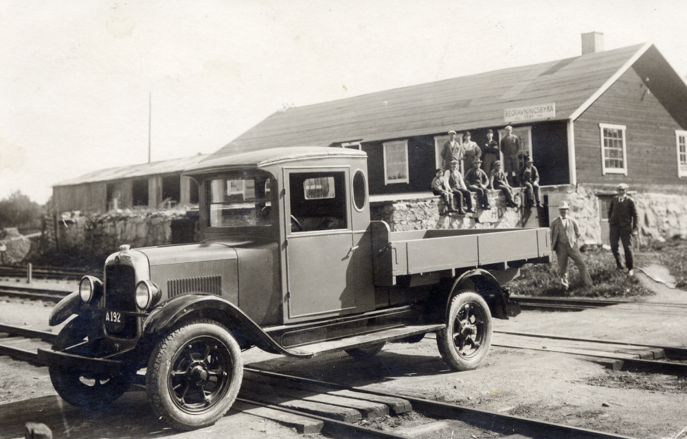 Vallentuna trävaruaffär grundades av S(igne) Linder 1921,  p g a tidigare skulder kunde brodern Sven inte få lån,men det var han som drev företaget. 1924 blev sågen kommanditbolaget Vallentuna trävaruaffär Linder och Lengquist.  Gottfrid Lengquist var Svens kusin.   På bilden står Sven Linder vid husets högra gavel,klädd i ljus kostym och ljus hatt.  Familjen Sven och Hedvig Linder flyttade till villa Svensborg på Allévägen så snart det stod klart.  Tidigare hade de bott i Villa Kullen väster om Väsby kvarn. Efter fem år överlät Sven Linder företaget till sina kompanjoner och arrenderade i stället Täby låd- och snickerifabrik.  Kopia av vykort poststämplat 19 augusti 1928 med text: Sänder härmed ett foto på fabriken samt på vår nya verksamhet (vi bygga nämligen bilhytter och flak till Hans Osterman).  Som synes har jag virkesmagasinet snart färdigt och kan nu på alvar ta emot beställningar. ... I veckan 25-30 ds flyttar vi till Täby.  Vid järnvägsövergången precis norr om Vallentuna järnvägsstation står en lastbil med hytt och flak.  På huset i bakgrunden sitter en skylt "Begravningsbyrå Täby"