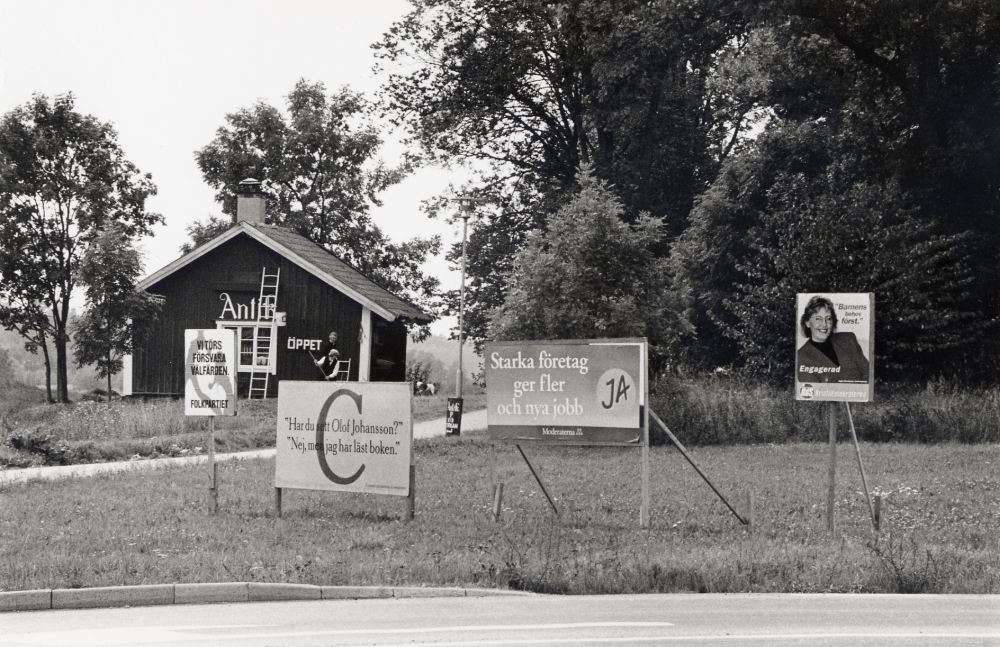 Valaffischer vid korsningen Väsbyvägen och Stockholmsvägen.  Valaffischerna är fr v Folkpartiet, Centern, Moderaterna och Kristdemokraterna.  I bakgrunden slår Ann-Marie och Berndt-Rune Jäverbrant gräset utanför Prästgårdens tvättstuga inför höstens försäljning i antikboden