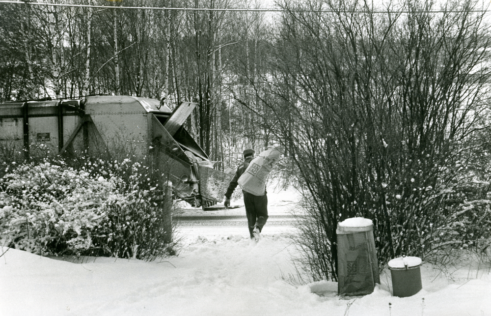 Sophämtning på Åbyholmsvägen. Sophämtaren bär en stor sopsäck fram till sopbilen, som har stannat till utanför tomten.

•Fotografiet ingår i bildserien ”Vardagsliv i Vallentuna kommun” av Rachael Gough-Azmier & Gunilla Blomé, Haga Studios HB.