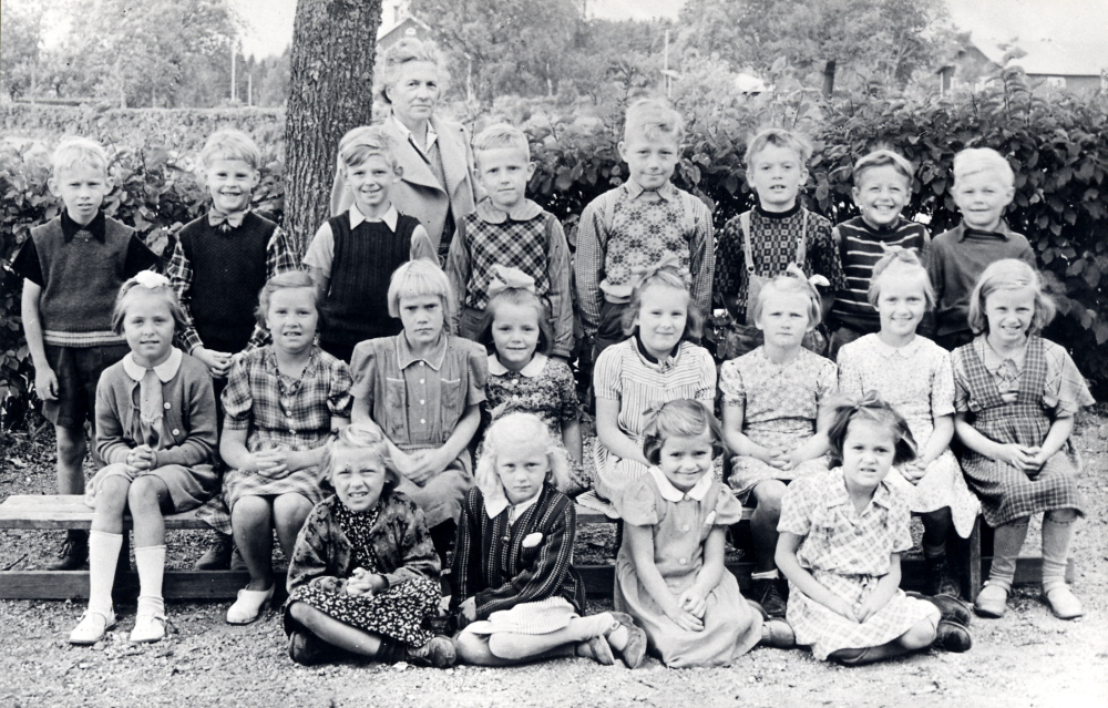 Lärarinnan Elna Sandgren tillsammans med skolbarnen i första klass vid Torsholma skola. Övre raden fr v: Bo Lundin, Lennart Söderström, Roger Qvarnström Tallen, Tavi Ränk, Lars-Erik Jansson Rolsta, Bjarne Zackariasson, Kurt Jansson Fredriksberg, Rolf-Arne Jansson Rolsta. Mellersta raden fr v: Ros-Marie Mattson Fågelsunda, Ulla Wredh Ragnhildsborg,  Maj-Britt Jansson, Irene Jansson Karlberg, Inga Johansson Tomthälla, Siv Nilsson, okänd, och Maj Johansson. Sittande fr v: Gunn-Marie Karlsson, Birgitta Berglund, Inga Helgöstam Helgö och Mona Sahlberg Rolsta