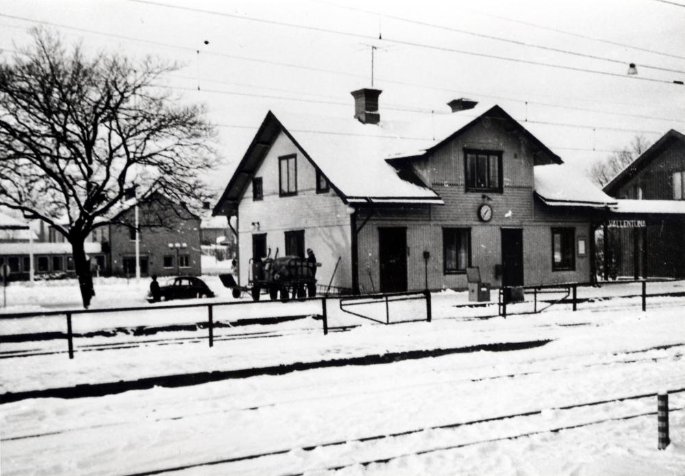 Vallentuna järnvägsstation och Vallentuna järnvägsrestaurang  t h. T v om stationshuset står en bagagekärra lastad med gods. I bakgrunden t v syns  Vallentuna kommunalhus. Framför stationshuset står en  Volkswagen  parkerad.