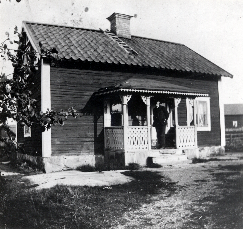 Carl Erik Norberg står på förstubron till Nannyberg i Rävsta. Snickerierna gjorde svärfadern Olsson, mjölnare på Rocksta kvarn år 1890. Stugan var från början garvarstuga i Brottby vid ån och flyttades tilll sin nuvarande plats 1890. I bakgrunden en av Rävstas ladugårdar. Carl Erik Norberg var fjärdingman