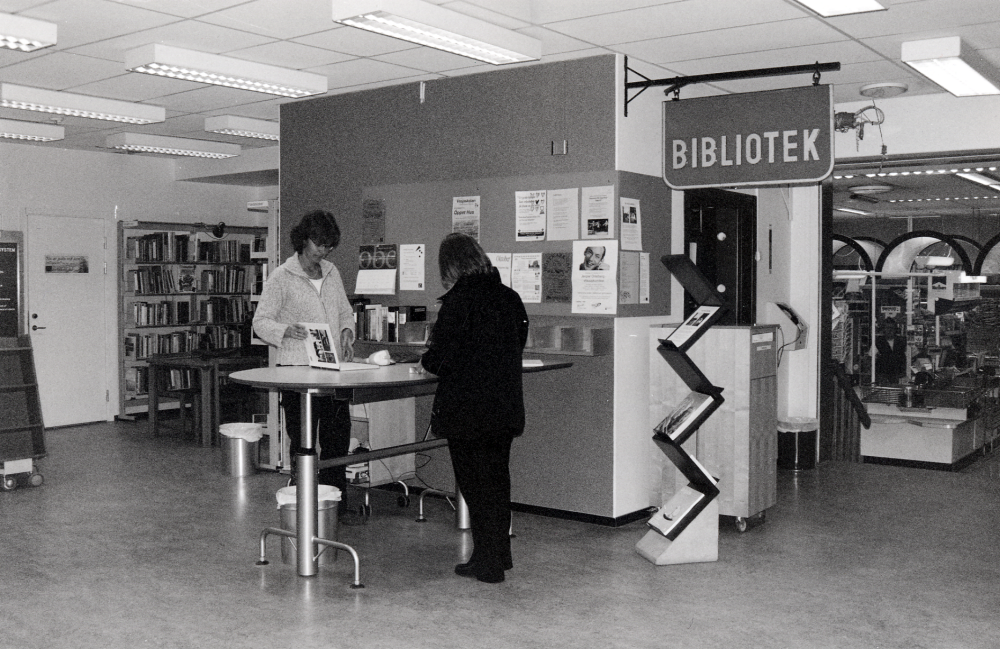 Karby bibliotek i samverkan med ICA Brottbyhallen vid Karby torg.  Biblioteket flyttade från Tallstigen 1994 och öppnade här i postens gamla lokaler.  Från början drevs biblioteket, posten och ICA i samverkan.  Posten flyttade efter några år till Bilisten vid Brottby torg.  2003 fräschades bibliotekslokalen upp med bl a ny färgsättning och nytt arbetsbord.  T h filialföreståndare Carina Schinkler och t v bibliotekarie Anna Bergström på besök från Vallentuna bibliotek.  Den gamla biblioteksskylten fanns från början i Össeby folkbibliotek i kyrkskolan i Lilla Garn.  I bakgrunden syns ICA-hallens affärslokal med trappa upp till biblioteket