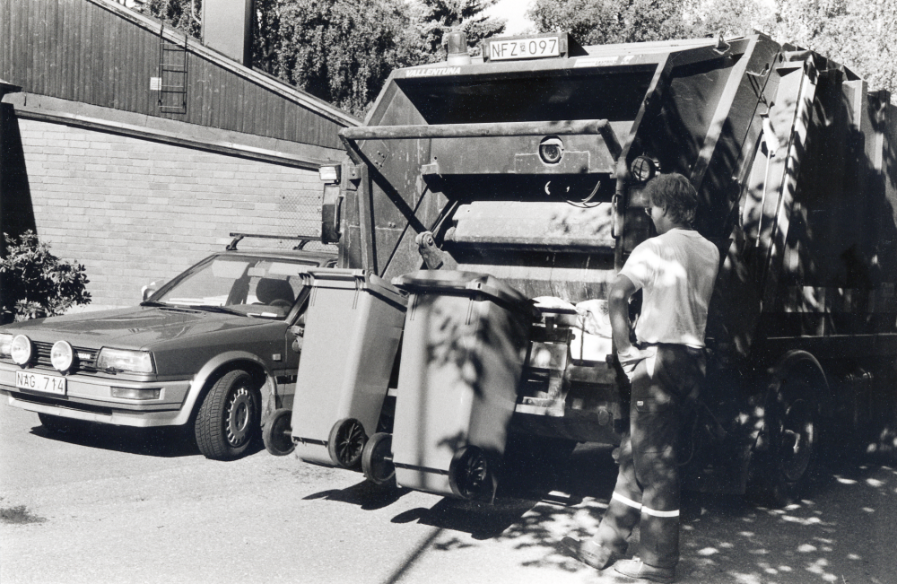 En av Ragn-Sells bilar tömmer soporna  i villaområdet Ekliden, Bällsta. Villaägaren kör själv ut sin soptunna till farbar bilväg.  Tunnan är försedd med hjul och sopbilen har griparmar eller hylla med stödram som på bilden.  Armen lyfter upp tunnan,  tömmer den i sopbilen och ställer sedan ner den tomma tunnan på plats igen.  Sopgubbarna slipper alla tunga lyft