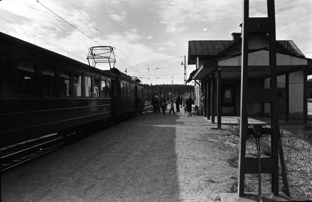 Ett tåg har stannat till vid Vallentuna station. Några resenärer kliver på. Stationshuset syns till höger.

•Fotografiet ingår i bildserien ”Vardagsliv i Vallentuna kommun” av Rachael Gough-Azmier & Gunilla Blomé, Haga Studios HB.