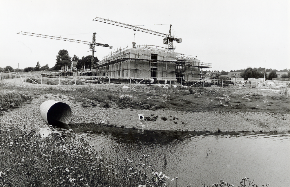 Vallentuna sjukhem och distriktläkarmottagning byggs vid korsningen Åby gata och Industrivägen. Här ses den blivande läkarmottagnigen från söder. Vattnet från Ormstaån rinner ur ett rör. T v syns en av tegelbruksvillorna och t h skymtar bebygglsen på Åby gärde. I förgrunden utloppet från Ormstaån