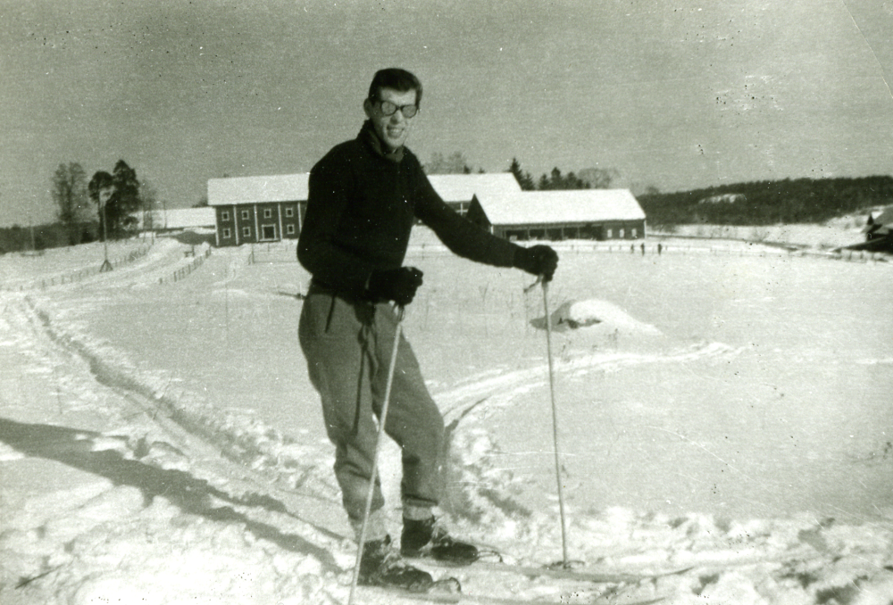 Joseph Spibey från London prövar skidåkning under ett besök i Kårsta. Det är första gången han står på skidor. I bakgrunden syns ekonomibyggnader på Backa gård. Vinterbild.