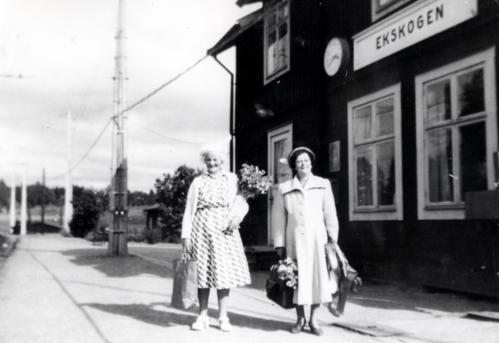 Ekskogens järnvägsstation. På plattformen framför stationshuset står Inez Ekholm, som tillsamman med sin man Manfred ägde Ekskogens gård 1847-1965, samt Adina Lind, en fröken som bodde i Stockholm. Fotografen är är adoptivson till Inez och Manfred Ekholm