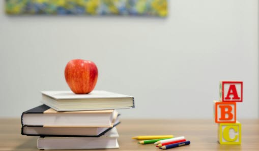 Bordsunderlag med staplade böcker med äpple vilandes högst upp. Intill ligger ett urval färgpennor och tre klossar på höjden i bokstäverna A, B och C.