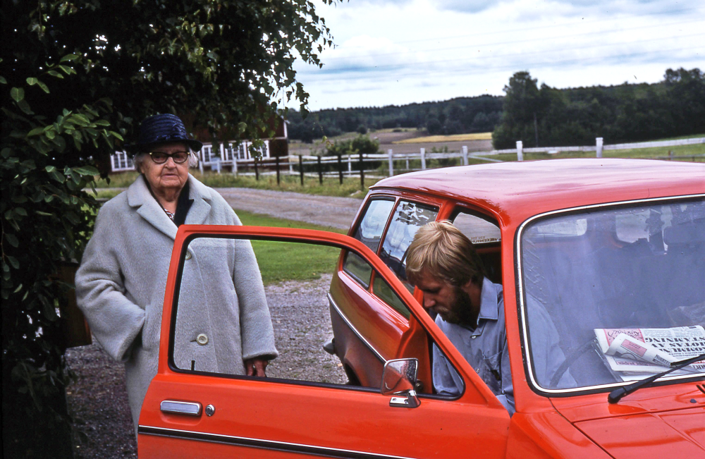 En arbetsdag med lantbrevbärare Mikael Mattsson. Lantbrevbäring i Rörby;  Adele Nilsson i Rörby, samtalar med lantbrevbärare Mikael Mattsson. Adele Nilssons man hette John Agaton Nilsson. Adele flyttade till Rörby 1961 och blev över 100 år gammal.  Ängarna ner mot Lövlund, numera Albyvägen syns i bakgrunden. Huset i bakgrunden är travstall till Albygård. 