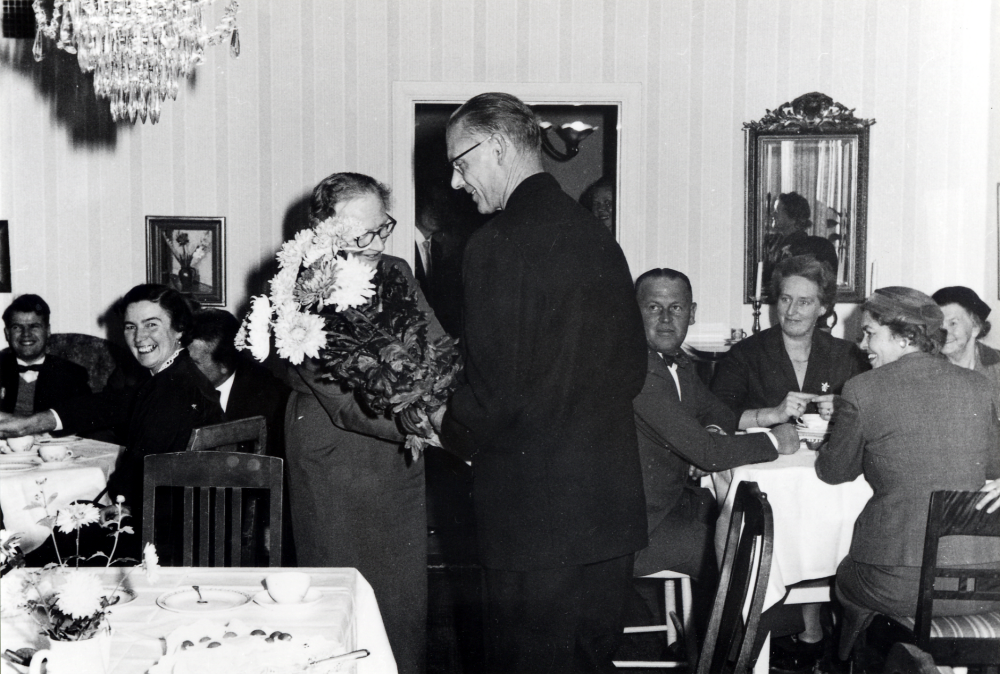 Prostinnan Ragnhild Vängby fyller 60 år och uppvaktas på högtidsdagen med blombukett  av Holger Hovmark.  I rummet sitter också  fr v okänd, Elna Hovmark, okänd, Göte Nilsson, Anna-Lisa Nilsson, Linnea Bengtsson och Elin Sundberg.  Ragnhild var gift med konraksprost Sven Vängby.  Förmodligen fotograferat i Vallentuna prästgård