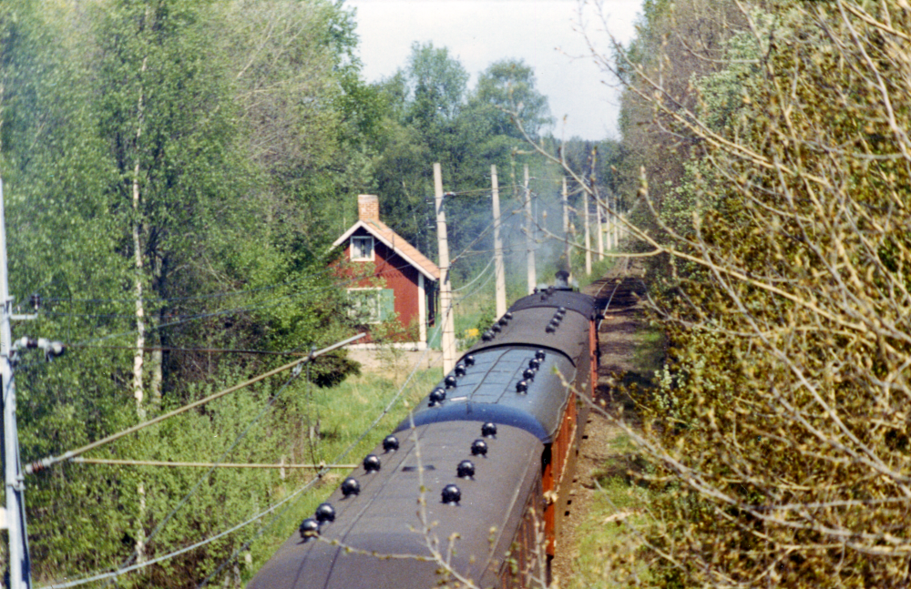 Norr om vägbron vid Veda ligger Grindvaktarstugan. Bilden kan vara tagen i samband med Roslagsbanans 100-årsjubileum 1985. Loket kan vara GP 3132 som även trafikerade Roslagsbanan vid enstaka tillfällen 1974-1977. Jämför med bild 7005.