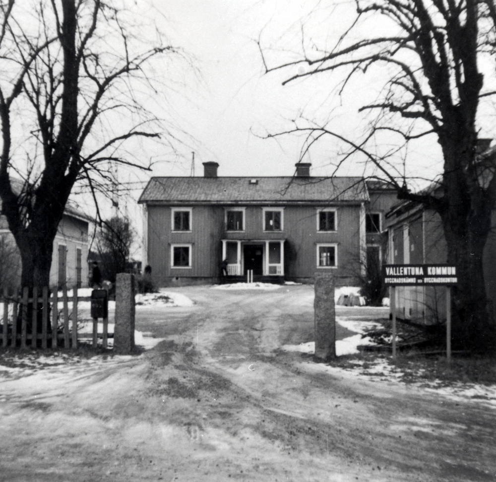 Väsby gård med flygelbyggnader sedda från söder med grindstolpar och vårdträd.  Grindstolparna och träden finns kvar 2003 och visar var gården en gång låg före rivningen 1971.  Denna bild togs i december 1970, precis före rivningen.  En skylt utanför grindarna visar att gården inhyser kommunen byggnadsnämnd samt byggnadskontor för de nya bostäderna i Sörgården