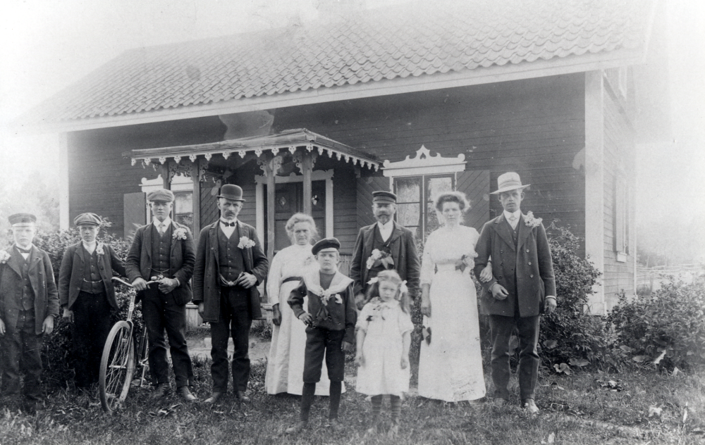 Utanför Nyborg i Össeby-Garn står fr v John Andersson 1894-1981, Hildur Andersson 1897-1964, Sven Andersson 1893-1977, Emil Karlsson gift med Georgina född Andersson, Matilda Andersson 1854-1931, August Andersson 1855-1930, Edith Forsberg 1886-1969 född Andersson samt Ivar Forsberg 1887-1954.  Barnen som står framför heter Helmer Karlsson född 1901 och är son till Georgina och Emil Karlsson samt Gertrud Johansson född 1905  och dotter till Tekla Johansson född Andersson