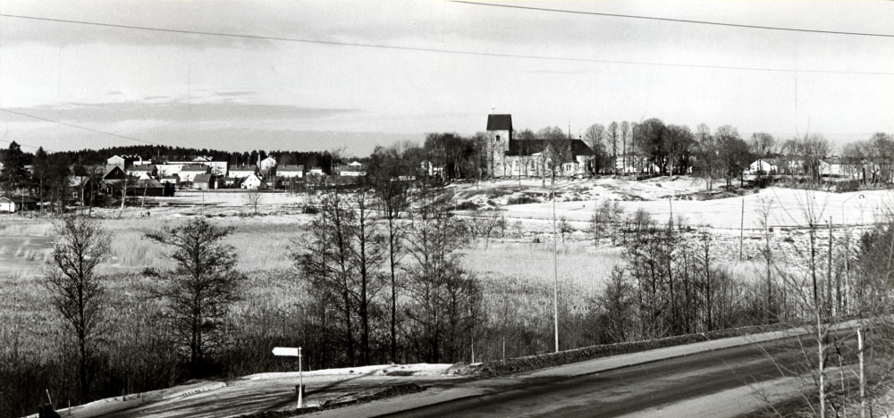Utsiktsbild över Vallentuna kyrka sedd från Bällsta. I förgrunden kan man skönja den gamla strandlinjen för Vallentunasjön ovanför tuvor och vass. Längst t v fanns på 1910-talet en gammal fiskarstuga som ägdes av fiskare Lejholm
