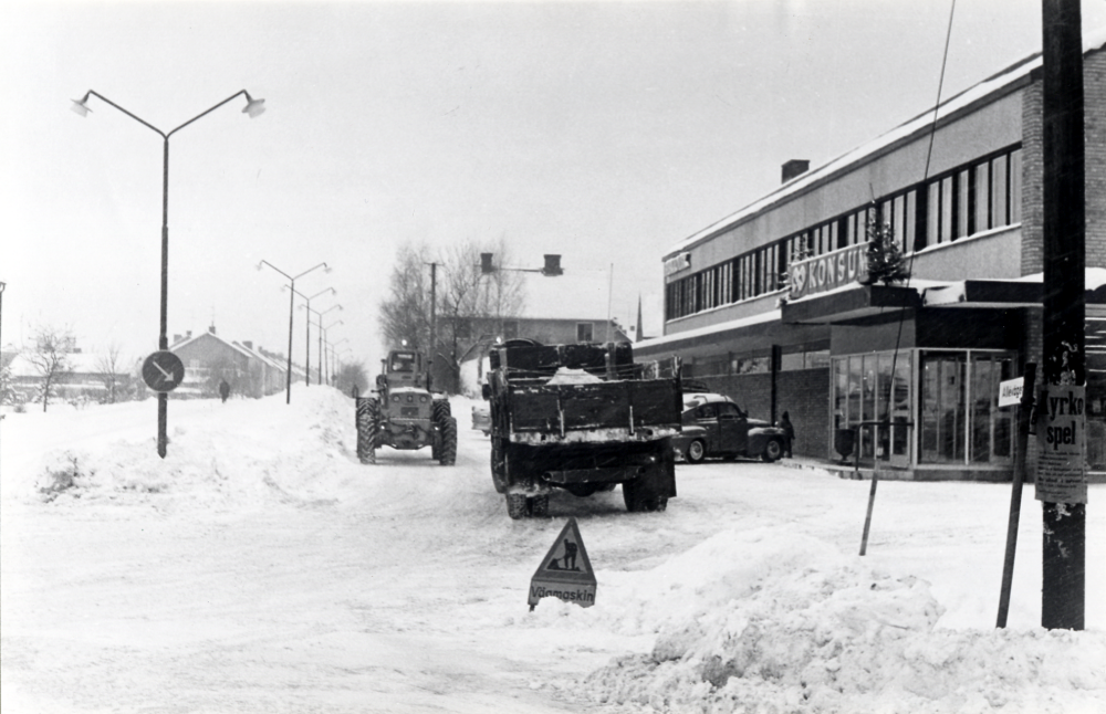 Snöröjning utanför Konsum på Allévägen med skoplastare och lastbil.  Julgranar står vid Konsums entré. På stolpen t h sitter en affisch om Kyrkospelet.