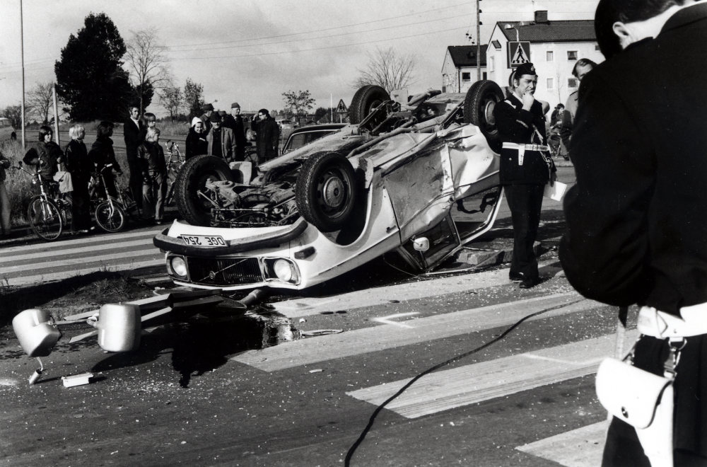 Trafikolycka i korsningen Stockholmsvägen och Väsbyvägen.  En ljus Volvo ligger på taket över trafikmärken och refug  på Stockholmsvägen.  I bildens mitt står Vallentunas  polischef Gunnar Söderquist.  I bakgrundens syns bostadshusen vid Centralvägen