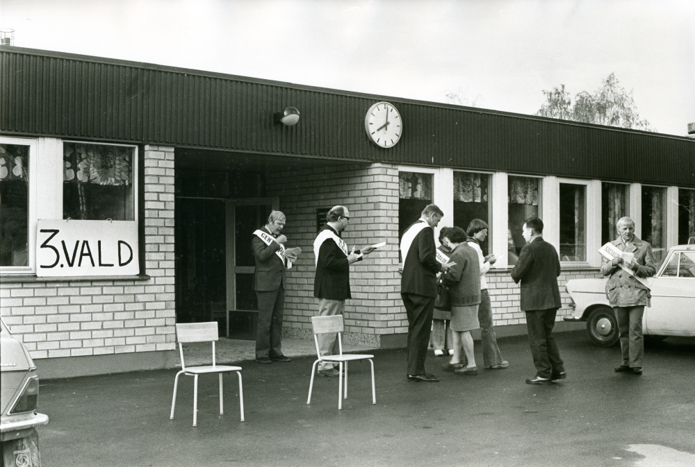 Klockan på fasaden har passerat 8 och vallokalen i Ormstaskolan har öppnat. Väljare strömmar till. Partifunktionärer från olika partier står utanför entrén för att dela ut sina valsedlar.
•Fotografiet ingår i bildserien ”Vardagsliv i Vallentuna kommun” av Rachael Gough-Azmier & Gunilla Blomé, Haga Studios HB.