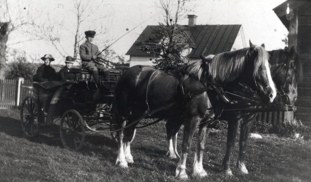Häst och vagn utanför Örsta gård i Markim.   Två hästar är spända framför vagnen.  På kuskbocken sitter Gustaf-Adolf Gustafsson.  I vagnen sitter fr v Olga Johansson och Alma Gustafsson