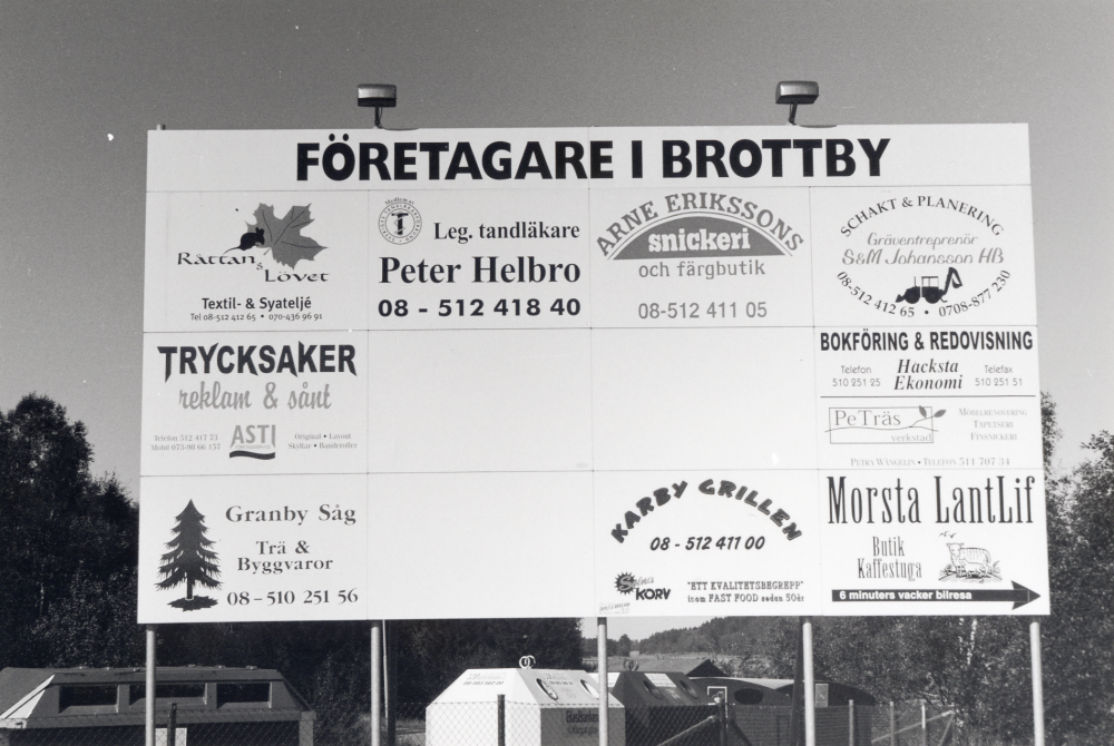 Företagare i Brottby.  Reklamskylt vid långtidsparkeringen nära Brottby trafikplats.  Bakom skymtar en återvinningsstation