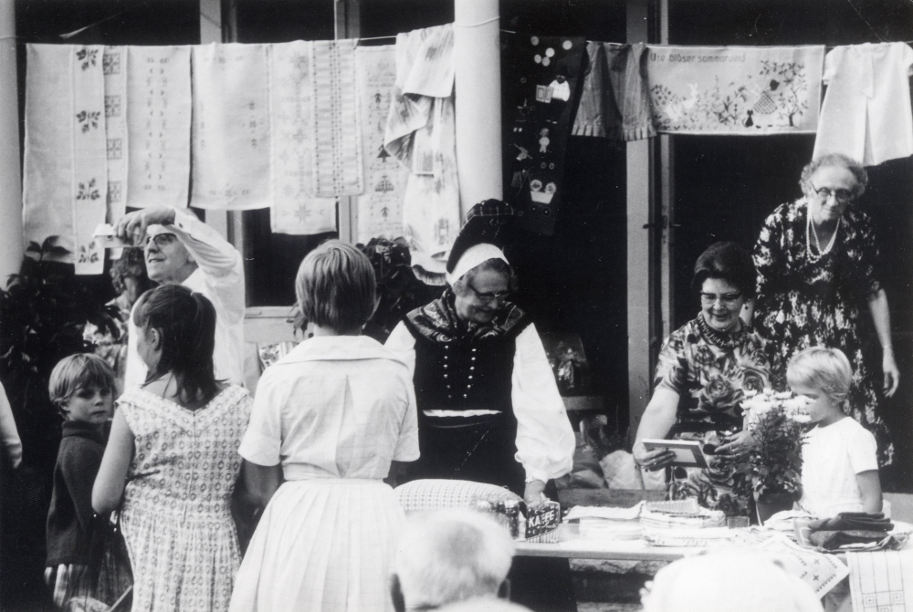 Traditionsenlig sommarauktion i Vallentuna prästgård då Kyrkliga syföreningens alster såldes. Utropare på bilden är lärarinnan Margit Oppermann i folkdräkt och kontraktsprost Sven Vängby i skjortärmar.  På verandan till prästgården hänger på snören handarbeten och dukar.  Damerna i blommiga klänningar fr v Thea Lembke och Ragnhild Vängby