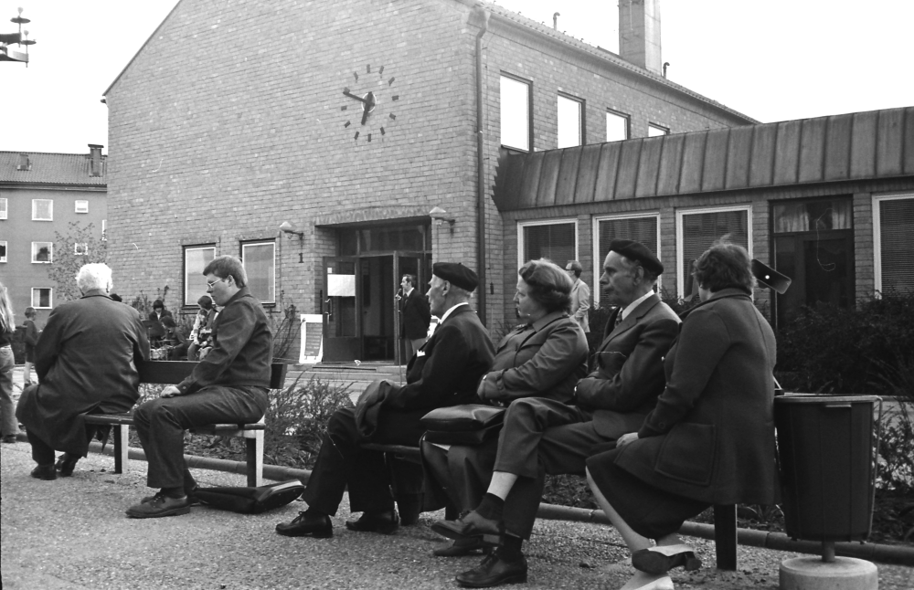 Några personer sitter på parkbänkarna utanför gamla kommunalhuset vid Tuna torg.
•Fotografiet ingår i bildserien ”Vardagsliv i Vallentuna kommun” av Rachael Gough-Azmier & Gunilla Blomé, Haga Studios HB.
