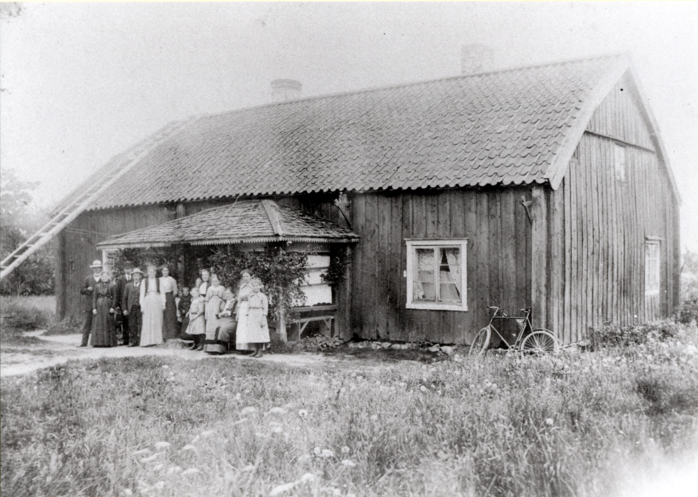 Gamla huset vid Örsta gård i Markim.  Huset revs 1915.  Fotografi från tiden 1890-1910.   Framför gården står fr v Carl och Ester Gustafsson, Alfred Jansson, okänd, okänd, okänd, Gustaf-Adolf Gustafsson, Vivan Jansson, Oskar Gustafsson, Maja Jansson samt Olga, Selma, Karin och Lilly Gustafsson