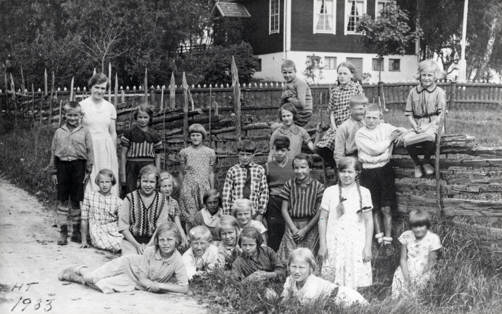 Lärarinnan Signe Jansson, 1944 gift Andersson tillsammans med skolbarnen utanför Åvasta skola. Liggande i gräset fr v: Doris Johansson, Erik Eliasson, Aina Lundh, Sanna Jansson, Ingegärd Eliasson och Britta Andersson. Andra raden stående Henry Larsson, sittande Anna-Lisa Wadström, Elin Johansson, Ulla Ekberg, Eivor Roos och Stina Andersson Bredvid fröken står t h Maja Jansson, Stina Karlsson, Lars Ekberg, Ingvar Fredriksson, Majsie Larsson och Märta Nilsson med flätor. Stående framför gärdesgården Inga Sandberg, Einar Eliasson och Bertil Grandin. På gärdsgården sitter Valle Johansson, Birgit Grandin och Dagny Sandberg