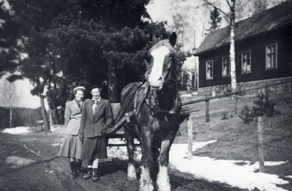Sursta kapell i västra Vallentuna syns t h.  I bildens mitt står två av evangelisterna som tjänstgjorde i kapellet, t v Ingegerd Johansson och t h Rosa Enstedt.  De lutar sig mot en vagn dragen av en häst.  Evangelisterna hjälpte ibland bönderna runtomkring