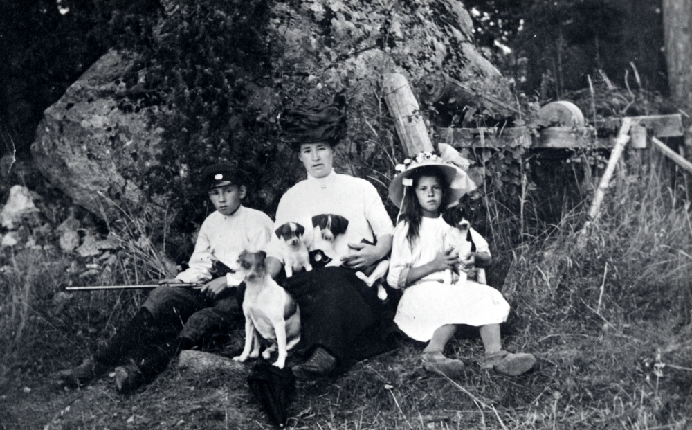 Mina Öberg   Davidsborg  tillsammans med barnen t v  David Öberg  och  Stina Öberg  i gröngräset. En  hund  Trissan sitter framför och Mina har två hundvalpar i knäet och Stina har en. I bakgrunden står en  slipsten  med vev